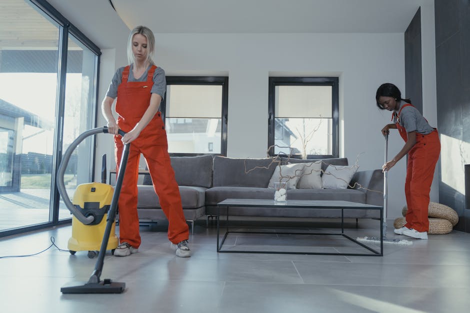 Close-up of three professional cleaners wearing grey shirts and orange overalls, holding vacuum cleaners, standing in a modern indoor setting with tiled flooring and a dark wall in the background. The