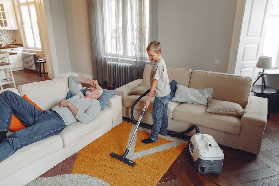 Close-up image of a compact vacuum cleaner with a transparent dust container filled with debris, connected to a flexible, patterned hose attached to a cleaning head on a pink carpeted floor. The vacuu