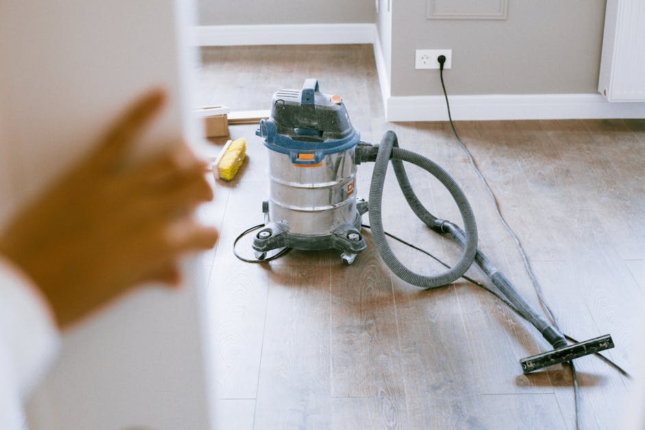 A close-up view of a professional cleaning vacuum cleaner placed on a wooden floor in a residential room. The vacuum, with a silver and blue body, has a black flexible hose connected to a metal wand and a cleaning head resting on the floor. In the background, a portion of a beige wall with a white electrical outlet and a radiator is visible, along with a window letting in natural light. A yellow cleaning brush is positioned near the vacuum. The scene depicts a thorough surface cleaning process, emphasizing the hygiene and maintenance involved in domestic cleaning services provided by Catford Cleaner, as seen in the context of deep cleaning for flats on Rushey Green SE6.