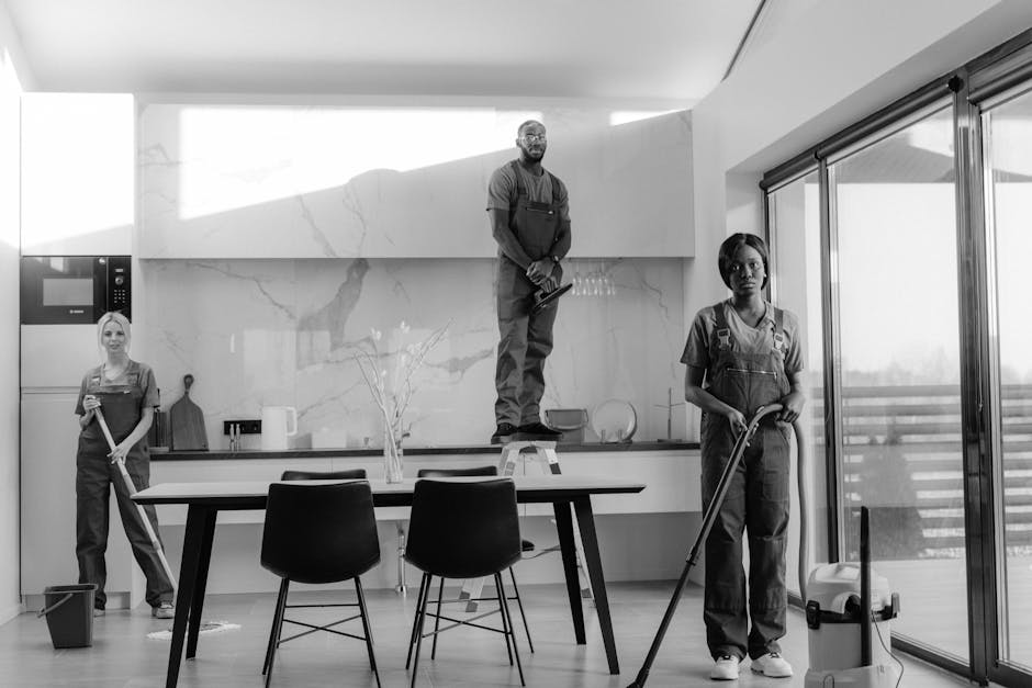 In a modern kitchen with a large marble backsplash and large sliding glass doors leading to an outdoor balcony, three professional cleaners from Catford Cleaner are engaged in surface cleaning. One cleaner stands on a step ladder in the center, wiping high up on the marble wall, while another on the right mops the smooth wooden floor, and a third on the left holds a mop and bucket, ready to clean the floor area. The room features a black dining table surrounded by three chairs, and a small vase with a plant sits on the table. The space is well-lit with natural light streaming through the glass doors, emphasizing the spotless and shiny surfaces. The cleaners are dressed in uniform, focusing on thorough deep cleaning and sanitization to ensure a hygienic, dust-free environment as part of expert domestic cleaning services from Catford Cleaner.