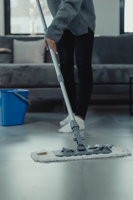A person mopping a smooth, light-colored tiled floor in a modern living room. The individual, dressed in dark pants and a grey sweater, is using a flat mop with a grey and white cloth attached to clean the surface. In the background, a grey sofa with beige cushions and a black side table are visible, along with a large window allowing natural light into the space. To the left, a blue bucket is placed on the floor, indicating a professional or thorough cleaning session. The area appears clean and well-maintained, showcasing surface cleaning and sanitisation typical of domestic cleaning services provided by Catford Cleaner near Catford Bridge Station.