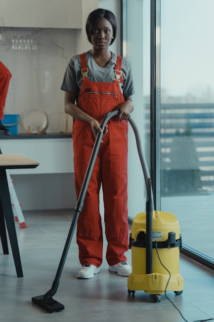 A person wearing a face mask, gloves, and an apron is engaged in surface cleaning at a shop with large glass windows, using a spray bottle and cloth to wipe the glass. The window frames are painted re