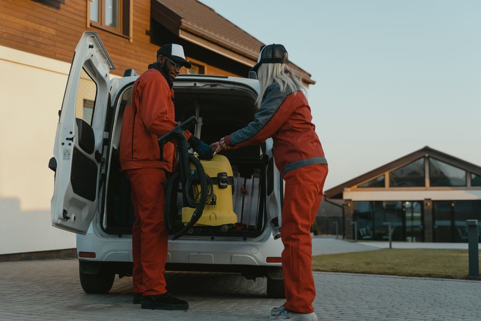 Two professional cleaners from Catford Cleaner, dressed in red work uniforms and protective caps, are preparing equipment from the back of a white van parked outside a residential building. One cleane